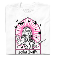 A folded white t-shirt with pink & black print depicting Dolly Parton with her arms outstretched. A stained glass frame is around her, with a pink sun behind her, black butterflies around her, and text at the bottom that says, "SAINT DOLLY."