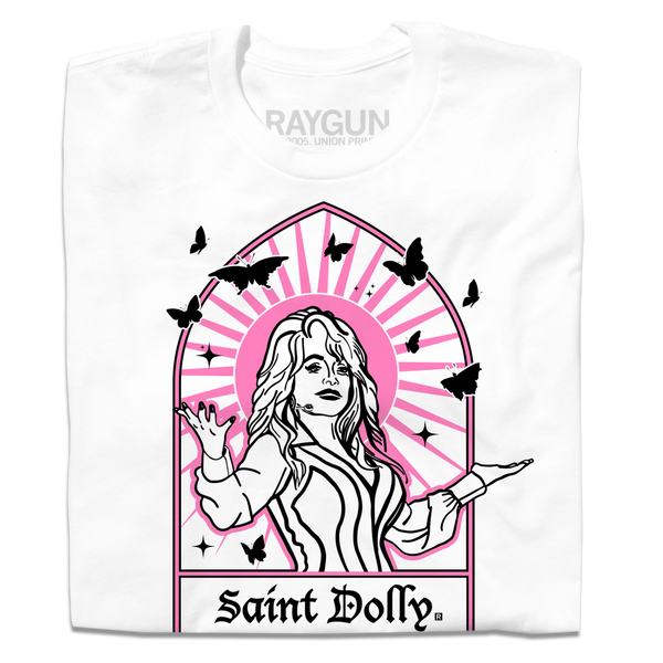 A folded white t-shirt with pink & black print depicting Dolly Parton with her arms outstretched. A stained glass frame is around her, with a pink sun behind her, black butterflies around her, and text at the bottom that says, 
