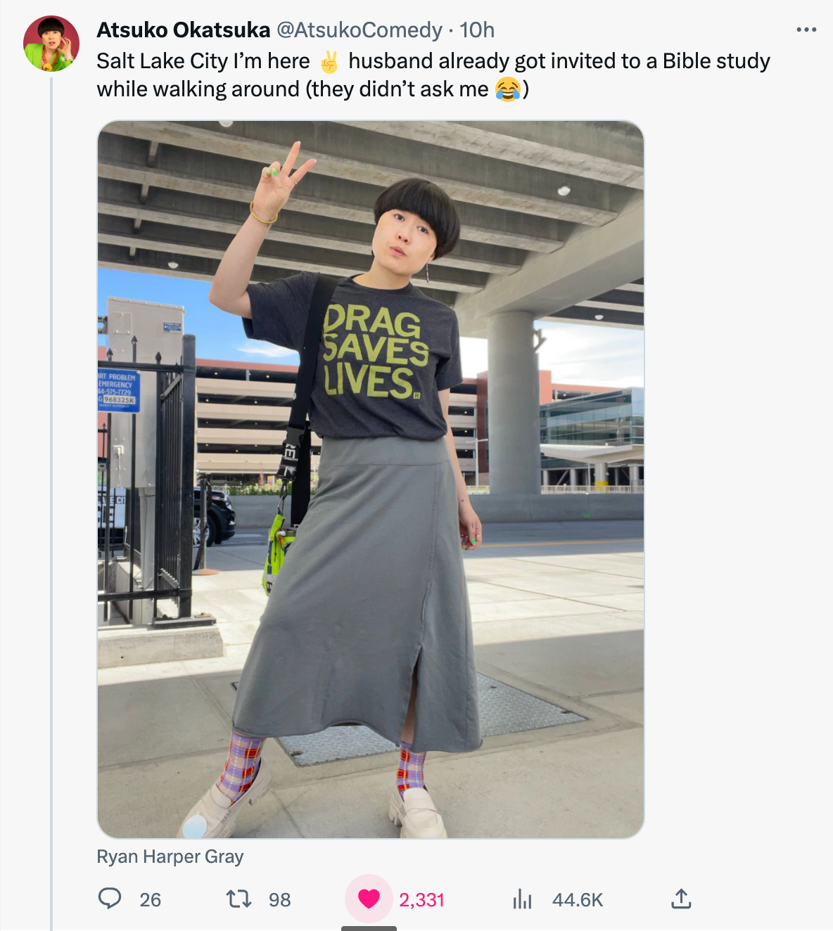A person wearing a charcoal DRAG SAVES LIVES t-shirt and a long grey skirt poses with a peace sign under a concrete overpass.