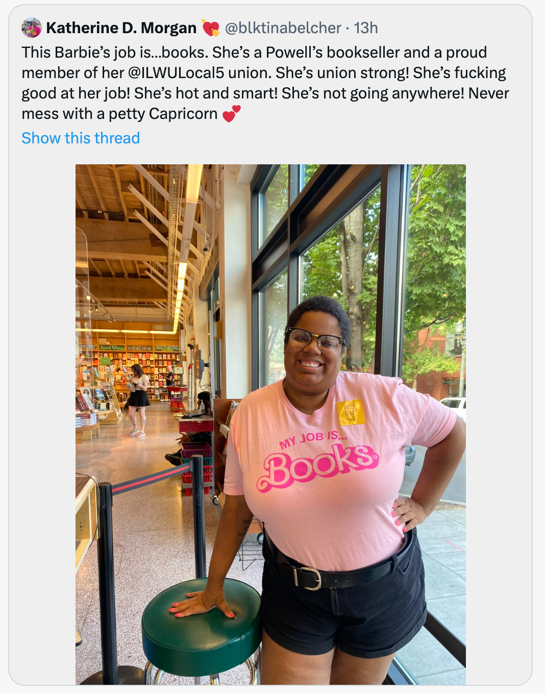 A smiling person wearing a sweatshop-free, pink t-shirt that says MY JOB IS... Books stands inside a bookstore.