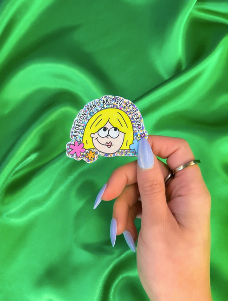 A hand with light blue nails holds a glittery vinyl sticker featuring a cartoon character and the text Abortion bans are so yesterday.