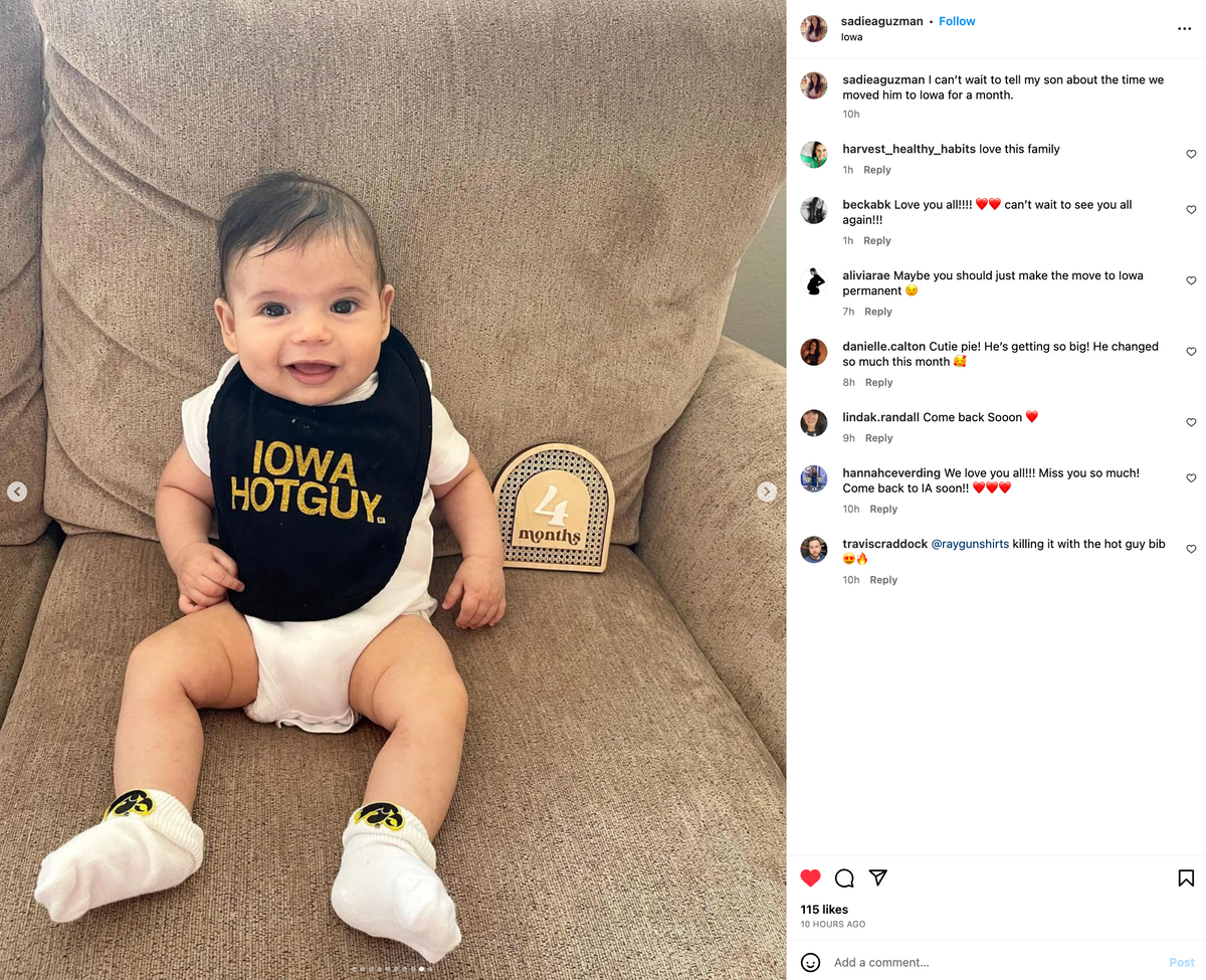 A baby wearing a RAYGUN Lil Iowa Hotguy bib sits on a chair next to a wooden sign that says 4 months.