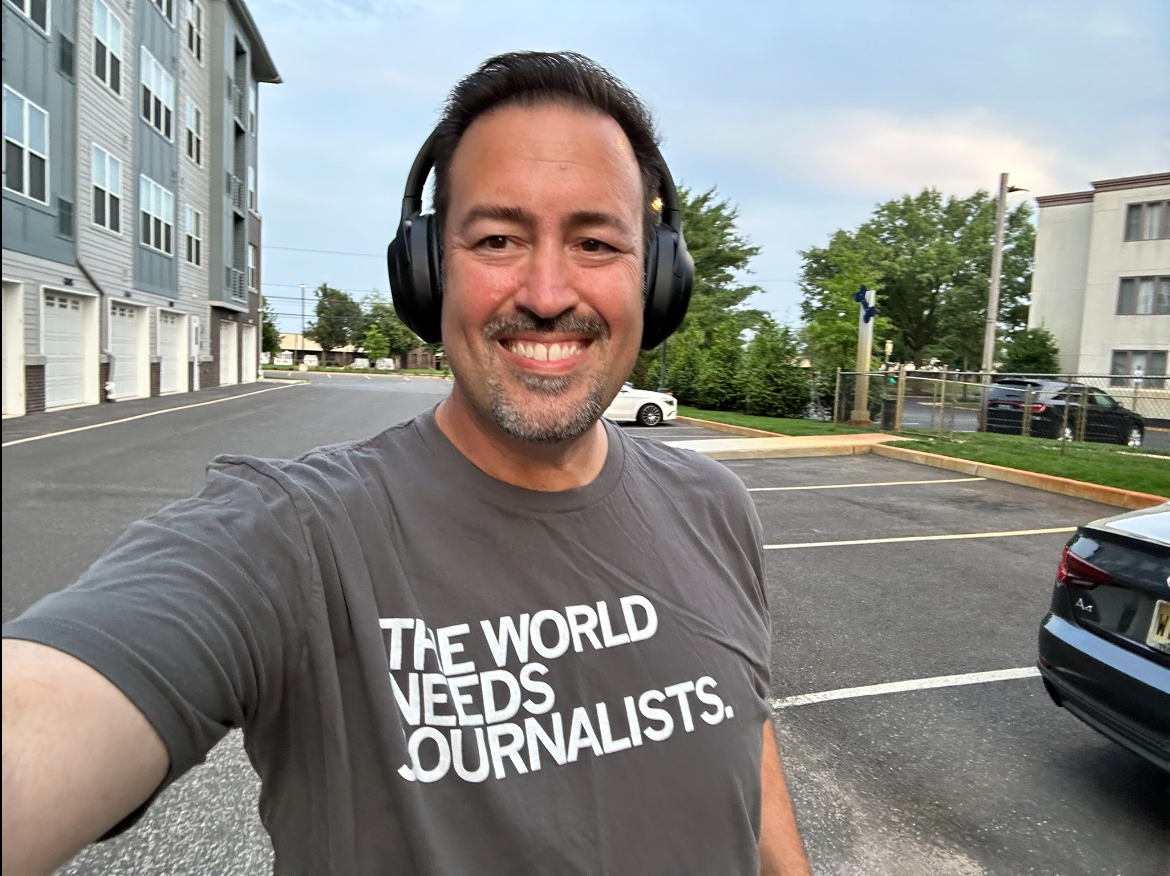 A man wearing black headphones and a gray RAYGUN t-shirt that says THE WORLD NEEDS JOURNALISTS smiles while taking a selfie in a parking lot.