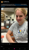 A woman wears a grey Heartland Pride t-shirt featuring a map of the United States with a heart in the center filled with the Progress Pride flag and the word HEARTLAND at the bottom.