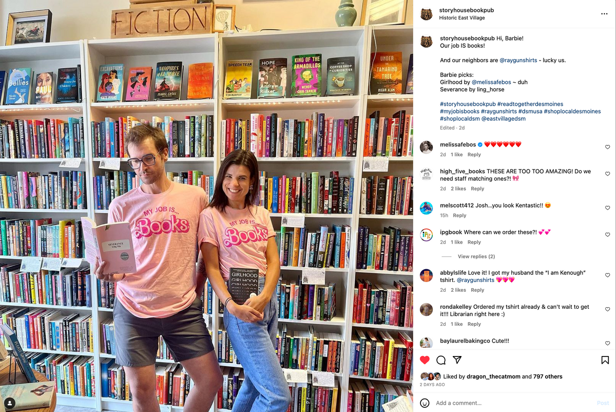 A man and a woman stand in a bookstore wearing matching pink books t-shirts that say MY JOB IS... BOOKS.