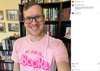 A man wearing glasses and a union made pink t-shirt that says MY JOB IS... BOOKS in a Barbie-style font, standing in front of a bookshelf.