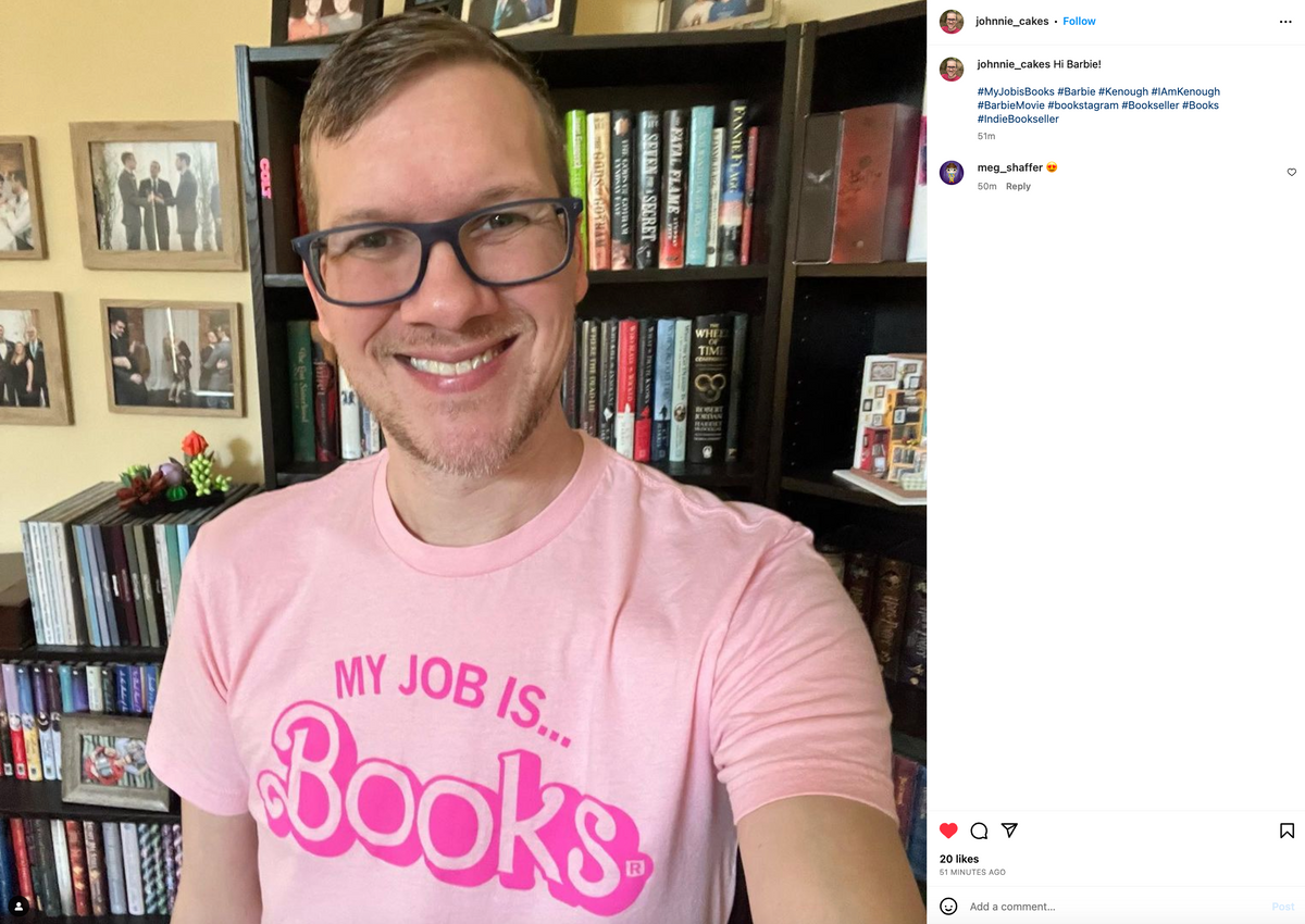 A man wearing glasses and a union made pink t-shirt that says MY JOB IS... BOOKS in a Barbie-style font, standing in front of a bookshelf.