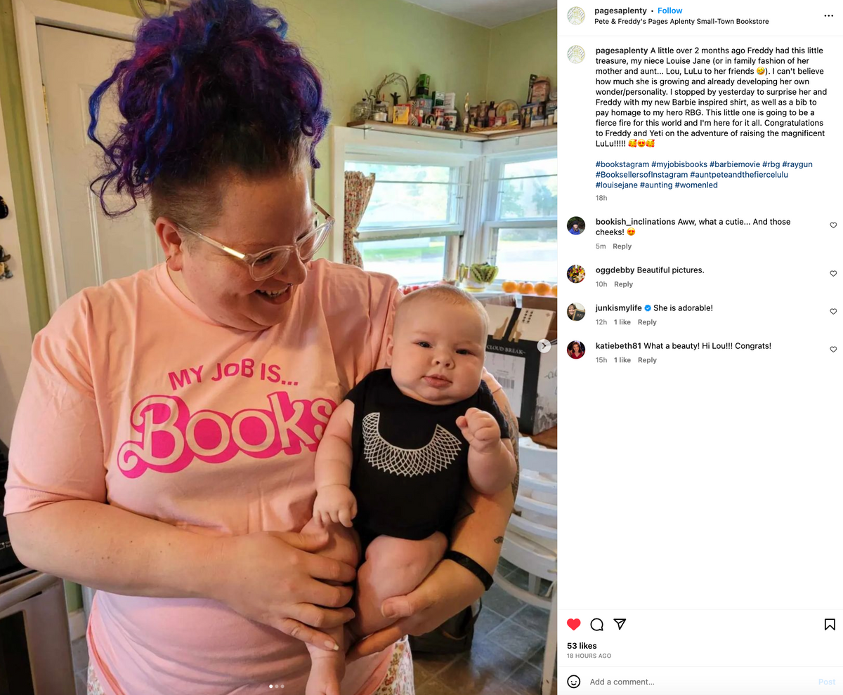 A person wearing a union made, pink My Job Is Books t-shirt holds a baby dressed in a black onesie printed with a white lace jabot.