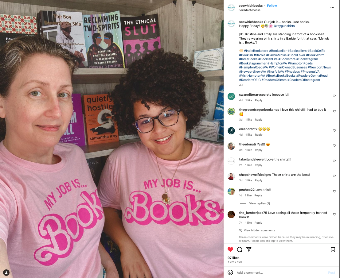 Two people standing in front of a bookshelf, both wearing matching union made, sweatshop free pink shirts that read My job is... BOOKS.