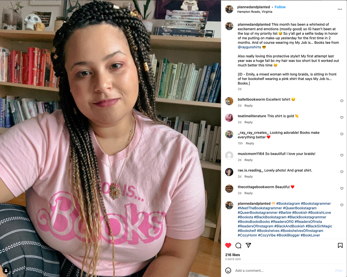 A woman with long braids wearing a pink My Job is... Books union-made T-shirt sits in front of a bookshelf.