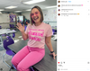 A teacher wearing pink glasses and a RAYGUN t-shirt that says This Barbie Reads Banned Books waves while sitting on a desk in a classroom.