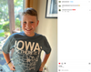 A smiling boy wearing an indigo t-shirt featuring a map of the United States with Iowa highlighted and the text IOWA OH, THERE IT IS.
