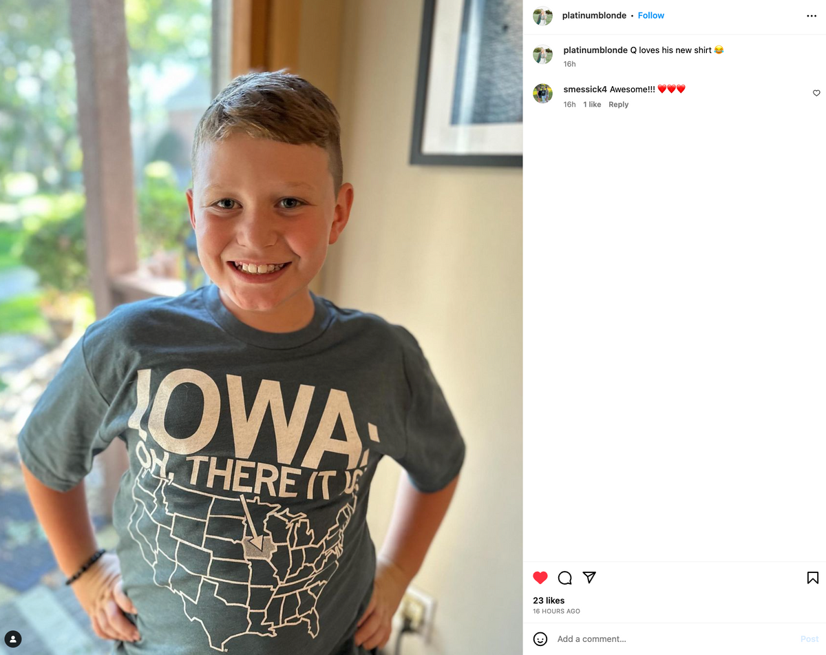 A smiling boy wearing an indigo t-shirt featuring a map of the United States with Iowa highlighted and the text IOWA OH, THERE IT IS.