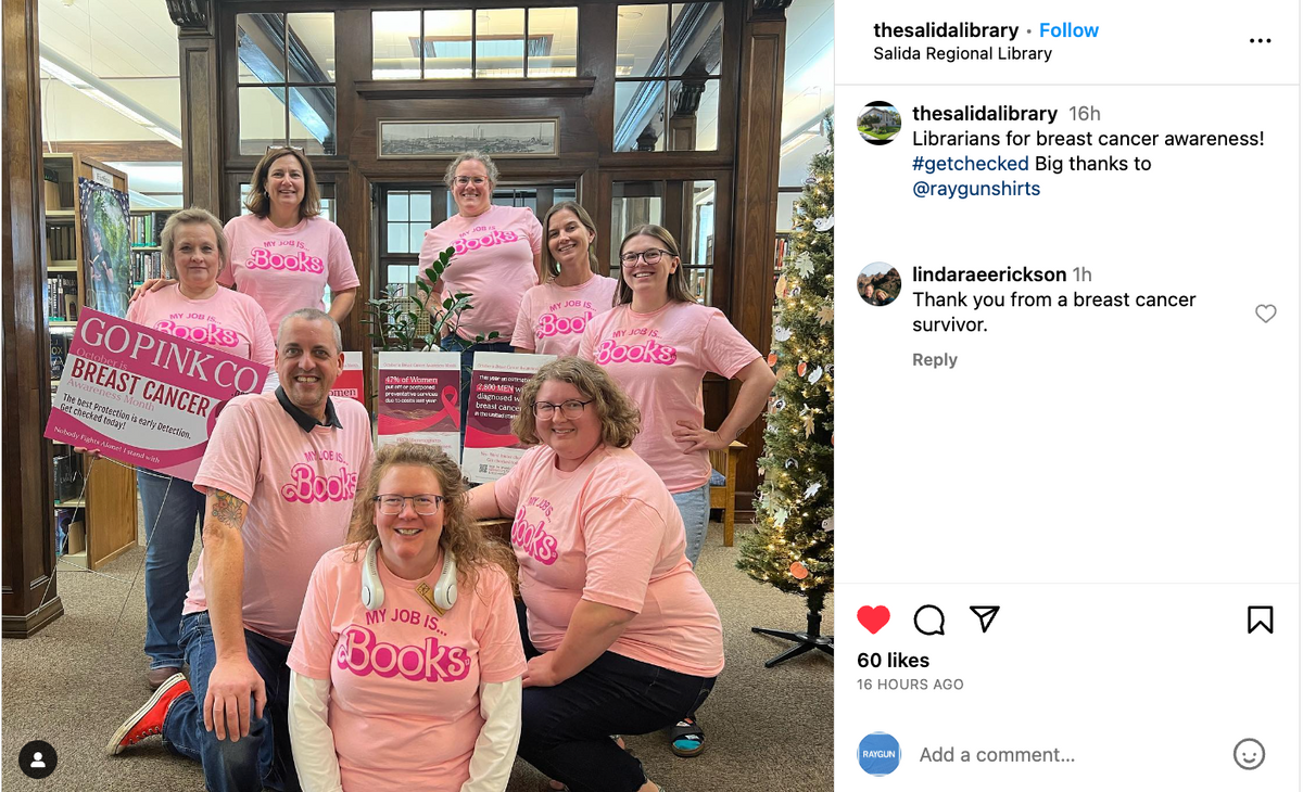 Eight librarians in union-made, pink My job is books t-shirts pose for a photo in a library, holding breast cancer awareness signs.