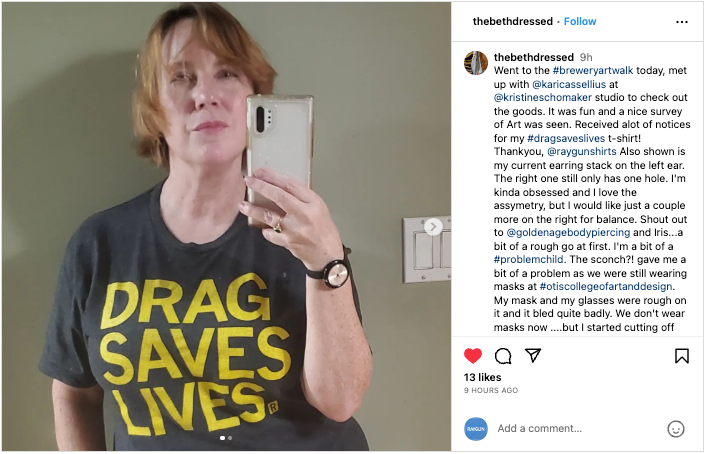 A woman taking a mirror selfie wearing a charcoal t-shirt with the yellow text DRAG SAVES LIVES.