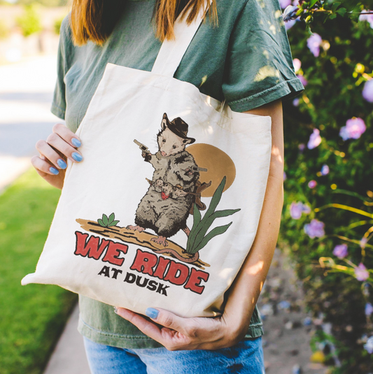 Canvas tote bag featuring a graphic of an opossum wearing a cowboy hat, holding two pistols, and carrying babies on its back, with the text We ride at dusk. This durable bag is perfect for daily use.