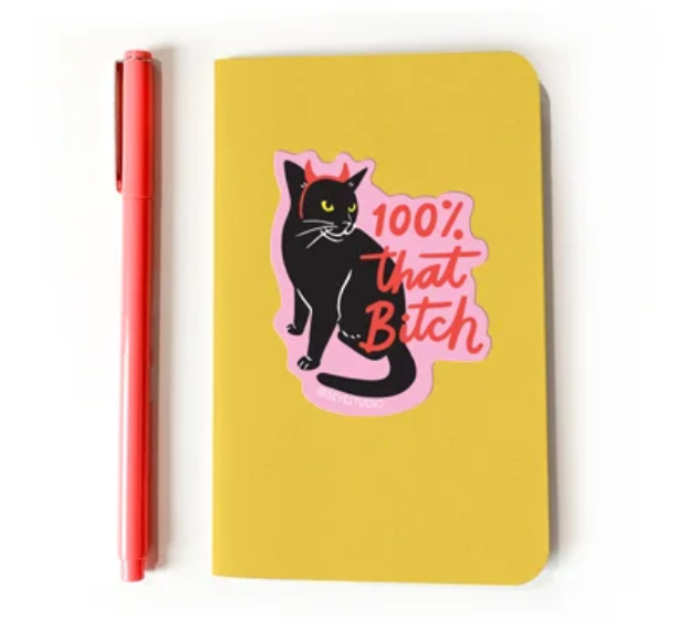 A yellow notebook featuring a high-quality sticker of a black cat wearing red devil horns with text that reads 100% that Bitch, placed next to a coral-colored pen.