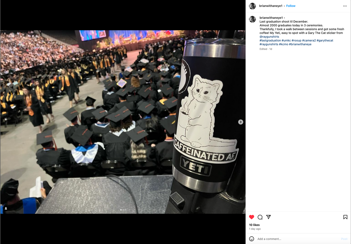 A black Yeti tumbler with a Caffeinated AF vinyl sticker of a cat holding a coffee mug, held in front of a graduation ceremony with many students in caps and gowns.