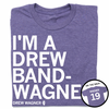 A folded heather purple t-shirt with left-aligned white text that reads "I'M A DREW BAND-WANGER" above smaller text that says, "DREW WAGNER" with a small outline of Illinois that has 19 in its center. A circle image in the bottom right corner shows the back of the t-shirt printed like a jersey with "WAGNER" above 19.