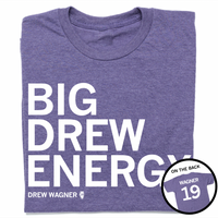 A folded heather purple t-shirt with left-aligned white text that reads "BIG DREW ENERGY" above smaller text that says, "DREW WAGNER" with a small outline of Illinois that has 19 in its center. A circle image in the bottom right corner shows the back of the t-shirt printed like a jersey with "WAGNER" above 19.