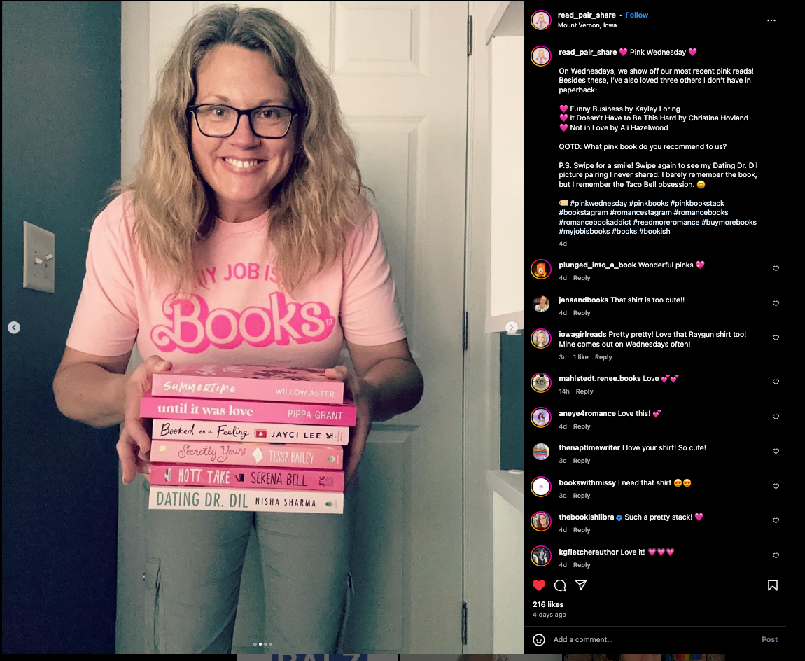 A smiling woman wearing glasses and a union-made, pink My job is books t-shirt holds a stack of five pink-themed romance novels.