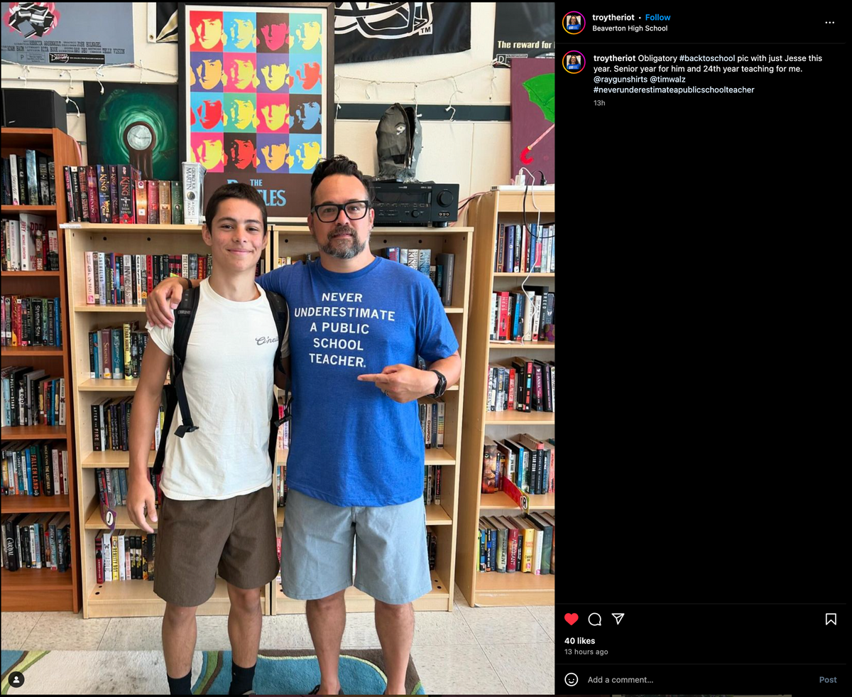 A high school student with a backpack stands with a teacher wearing a blue Never underestimate a public school teacher union-made shirt in front of classroom bookshelves.