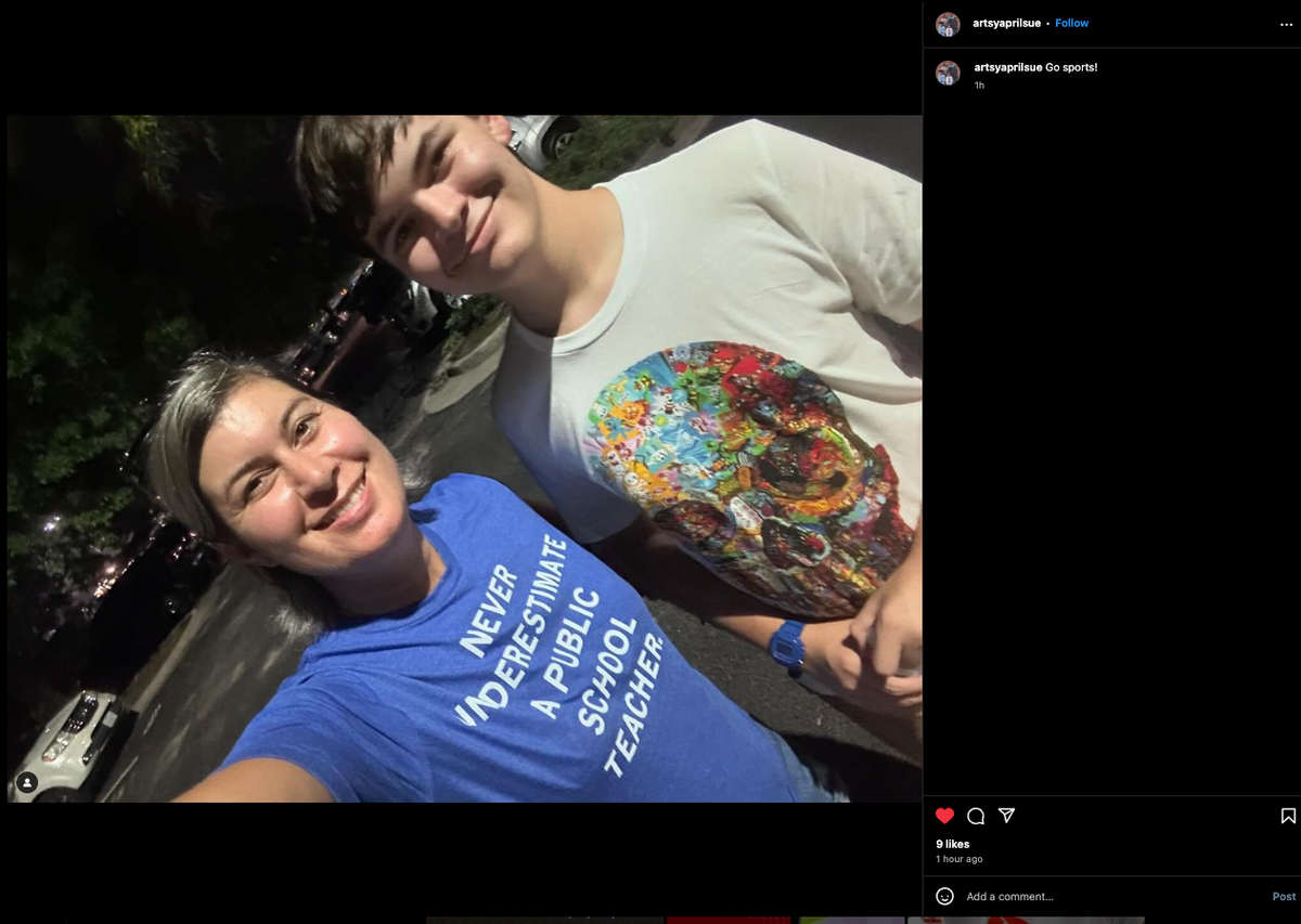 A woman wearing a blue teacher t-shirt that says Never underestimate a public school teacher and a young man in a white t-shirt with a graphic print pose for a selfie outdoors at night.