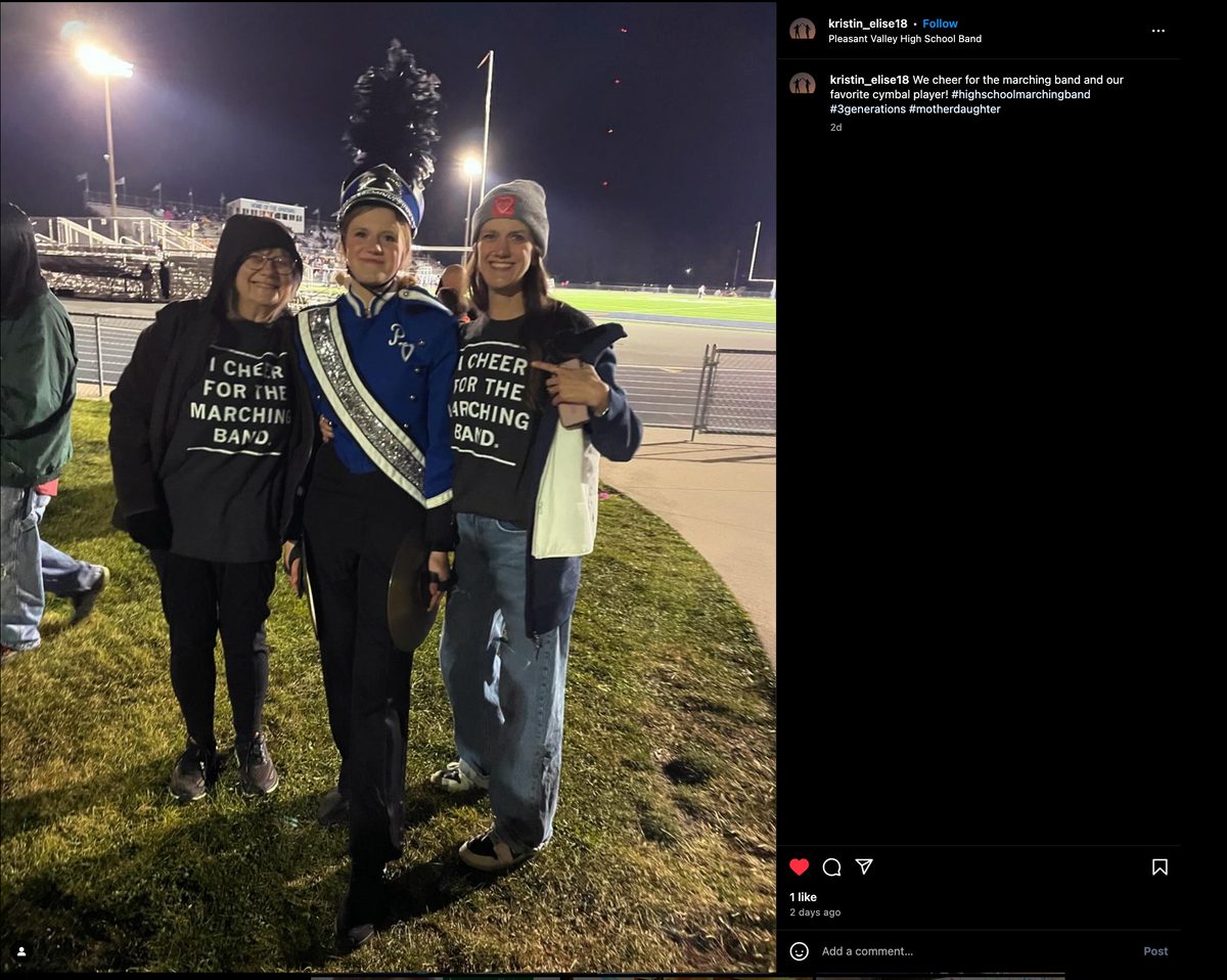 A marching band student in uniform stands between two women wearing matching charcoal t-shirts that read I cheer for the marching band at a nighttime football stadium.