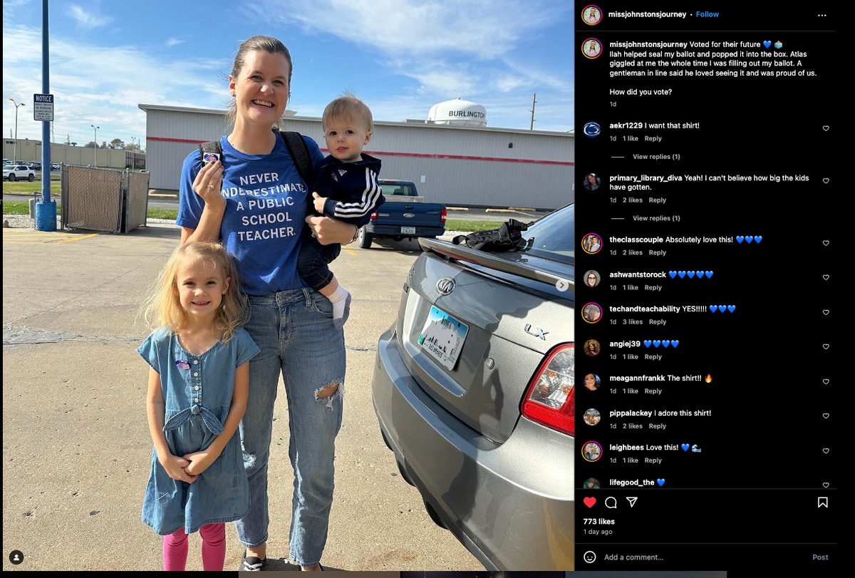 A woman wearing a t-shirt that reads Never underestimate a public school teacher stands in a parking lot holding a toddler, with a young girl standing beside her.