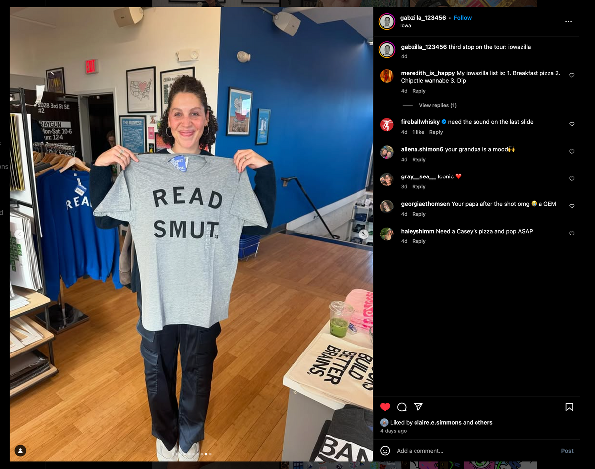 A person holding up a heather grey RAYGUN t-shirt with the words READ SMUT printed in black letters.