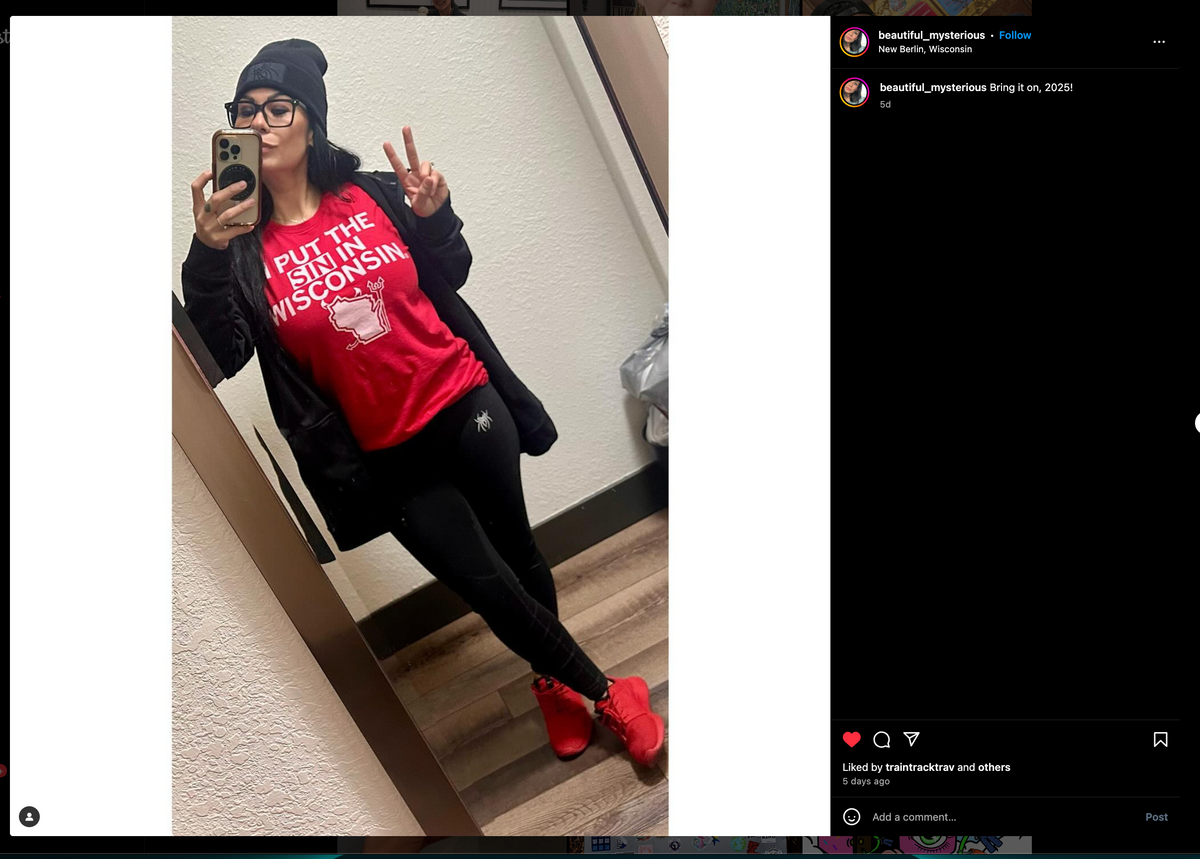 A woman in a black beanie, black zip-up hoodie, and a union-made red t-shirt that says I put the sin in Wisconsin takes a mirror selfie while making a peace sign.