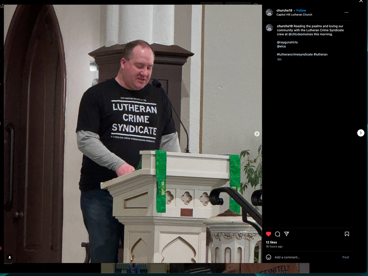 A man wearing a union made Lutheran Crime Syndicate t-shirt stands at a church lectern, reading from a document.