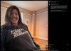 A woman smiles while wearing a Pigment Black Pullover Hoodie that says AMERICA NEEDS JOURNALISTS in white text.