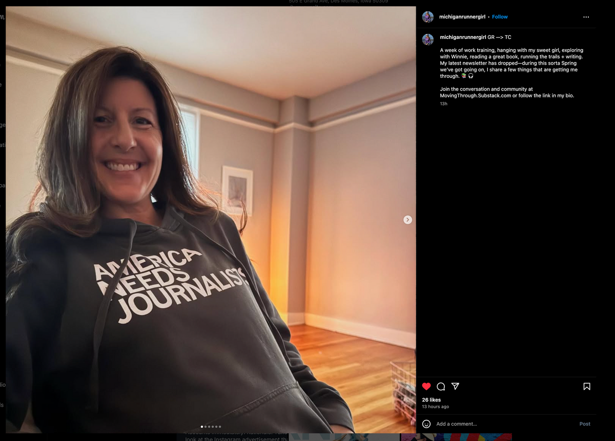 A woman smiles while wearing a Pigment Black Pullover Hoodie that says AMERICA NEEDS JOURNALISTS in white text.