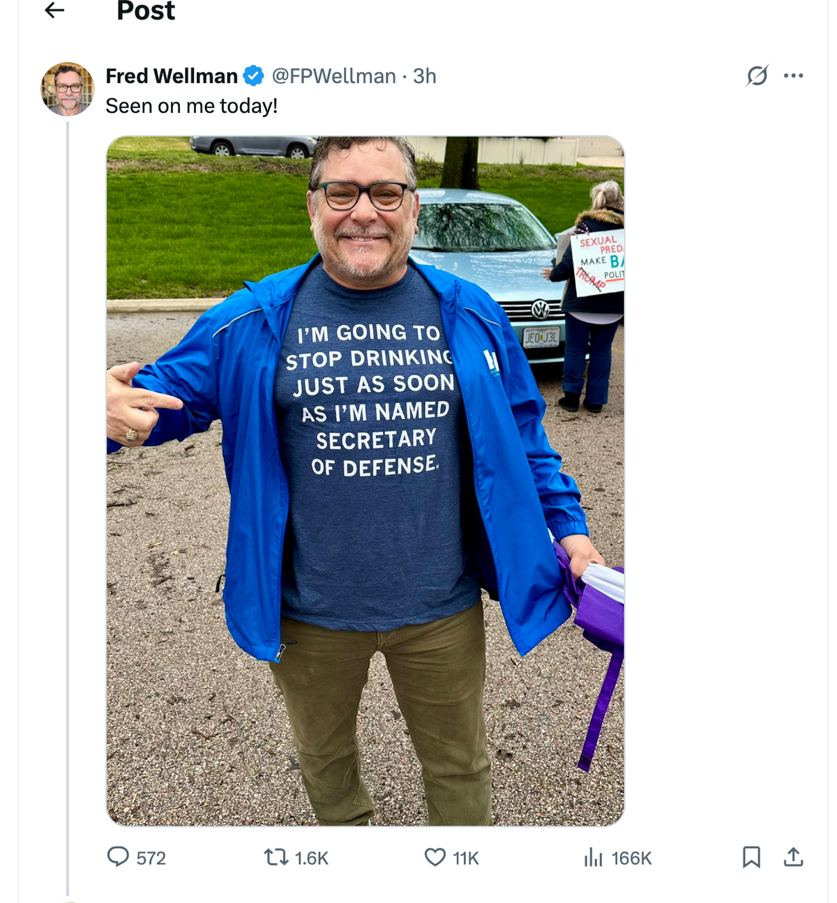 A smiling man wearing glasses and a blue jacket over a RAYGUN t-shirt that says, I’m going to stop drinking just as soon as I’m named Secretary of Defense.