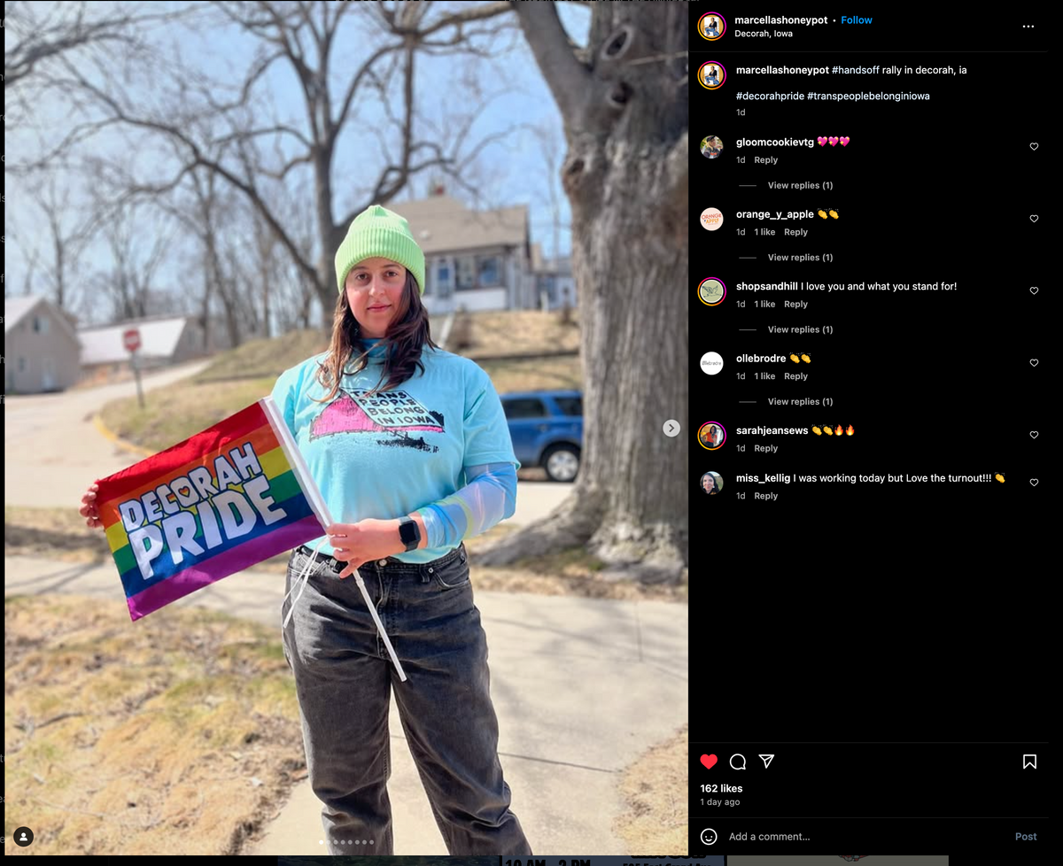 A person wearing a neon green beanie and a union made t-shirt that says Trans people belong in Iowa holds a small pride flag that reads Decorah Pride.