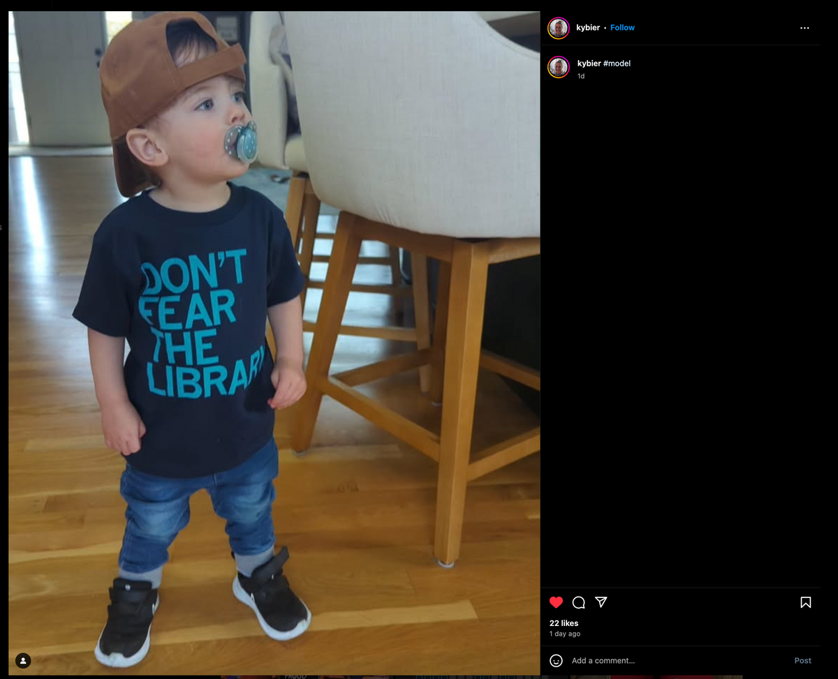 A toddler with a pacifier wears a brown baseball cap, sneakers, jeans, and a black ACLU shirt that says DONT FEAR THE LIBRARY.