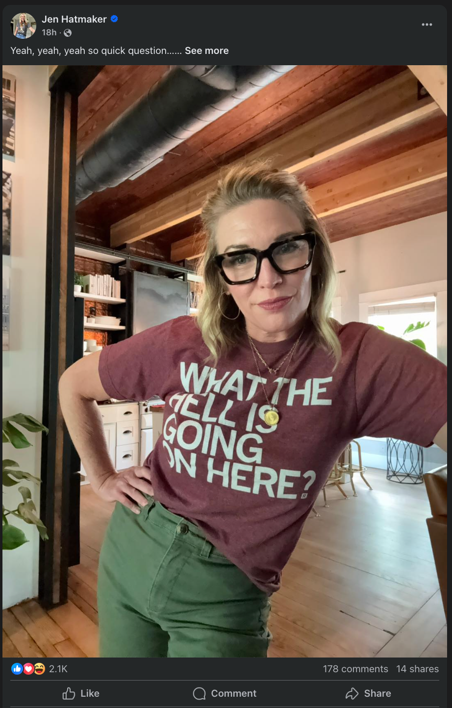 A woman wearing thick black-rimmed glasses and a maroon RAYGUN t-shirt that reads WHAT THE HELL IS GOING ON HERE? stands with her hand on her hip.