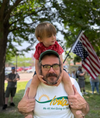 A man with glasses and a beard carries a young child on his shoulders outdoors near an American flag, representing the union labor families who rely on SNAP.