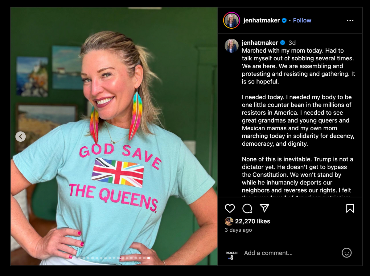 A woman wearing a heather purist blue t-shirt that says GOD SAVE THE QUEENS with a Union Jack icon and long, multicolored fringe earrings.