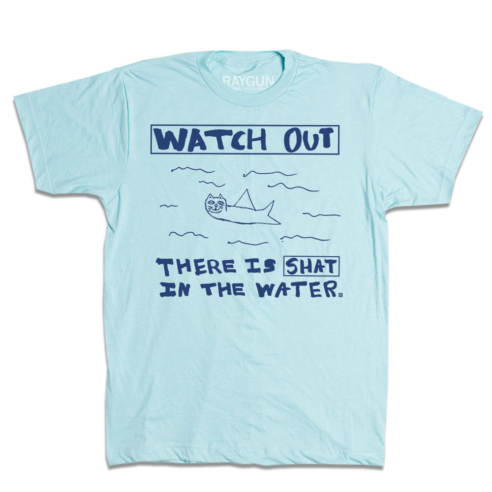 Kids size pastel blue t-shirt with royal blue print. A doodle of a shark/cat hybrid is in the center with text above and below that reads "Watch Out. There is a Shat in the water." This shirt was designed by a kid taking part in a contest at our Cedar Rapids store.
