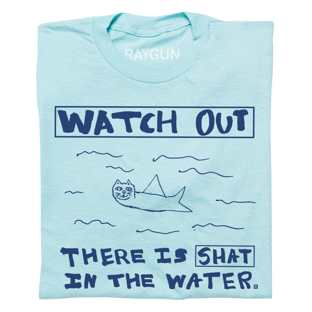 A folded pastel blue t-shirt with royal blue print. A doodle of a shark/cat hybrid is in the center with text above and below that reads "Watch Out. There is a Shat in the water." This shirt was designed by a kid taking part in a contest at our Cedar Rapids store.