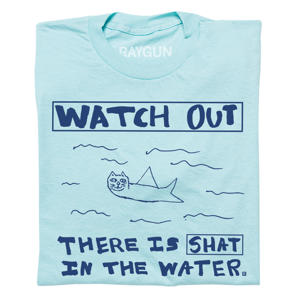 A folded pastel blue t-shirt with royal blue print. A doodle of a shark/cat hybrid is in the center with text above and below that reads 