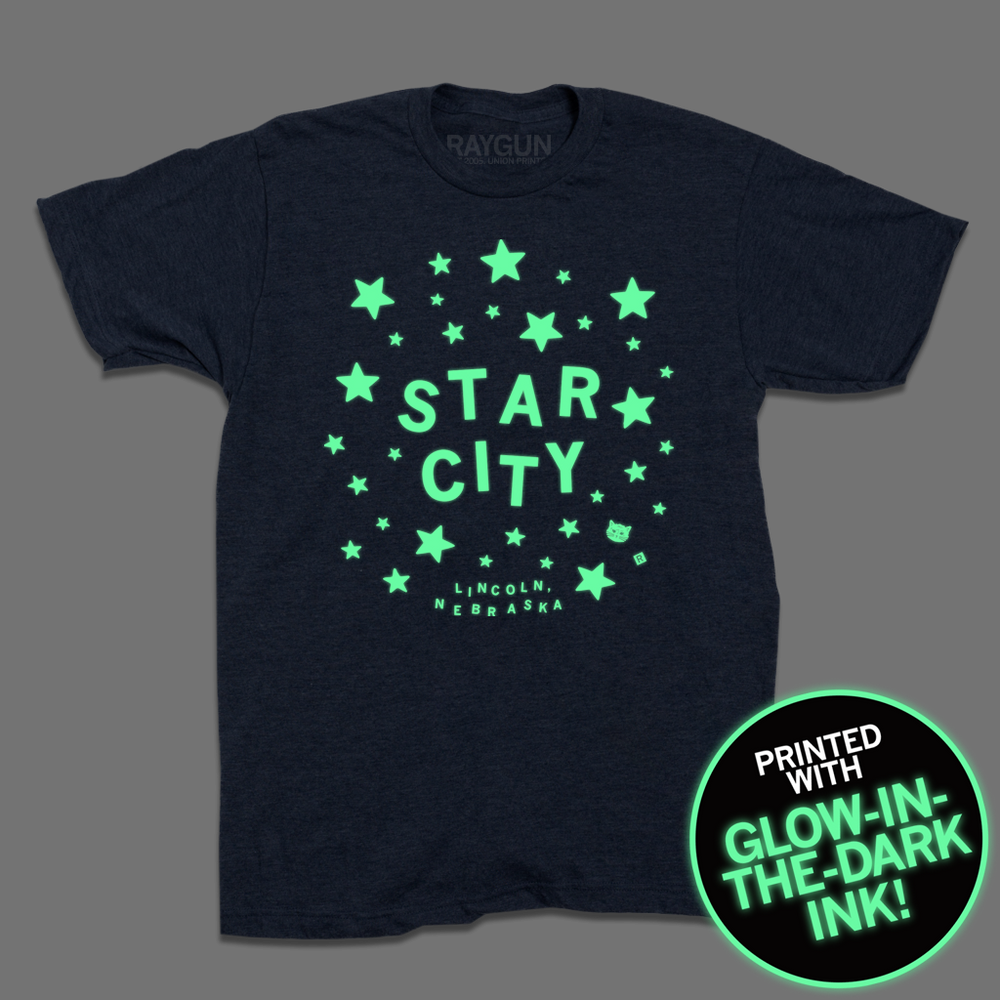 A heather denim t-shirt with glow in the dark ink that reads "Star City. Lincoln, Nebraska" in lit up text surrounded by lit up stars.