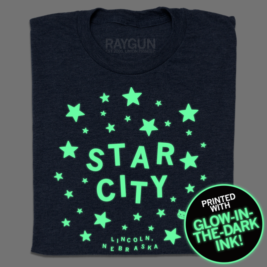 A folded heather navy t-shirt with glow in the dark ink that reads "Star City. Lincoln, Nebraska" in lit up text surrounded by lit up stars.