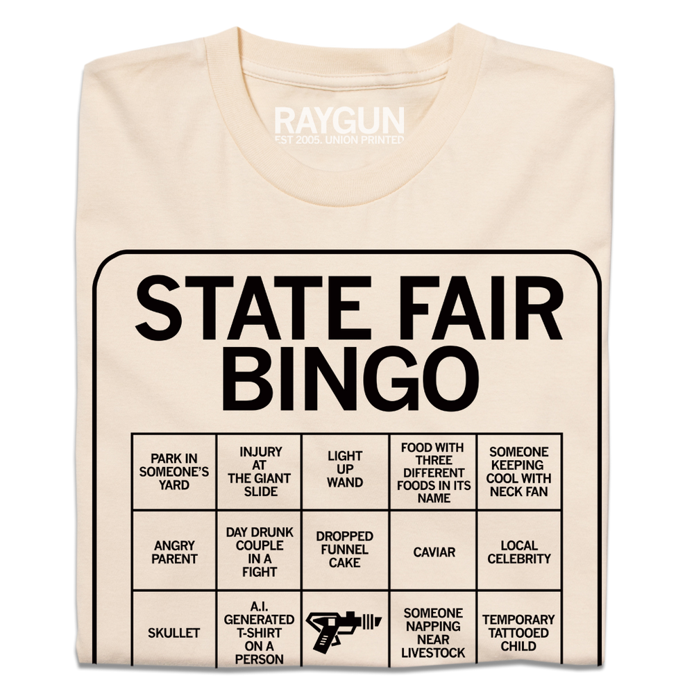 A folded off white shirt with black print depicting a bingo card. All caps text at the top of the card says, "State Fair Bingo" and the squares list things like "injury at the giant slide" and "someone napping near livestock."