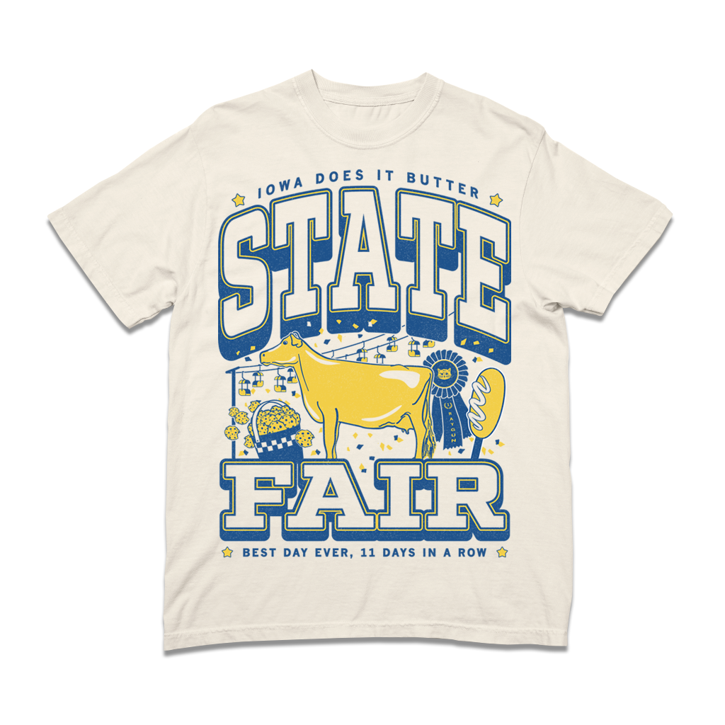 Cream-colored, union-made heavyweight t-shirt with an Iowa State Fair graphic that reads Iowa Does It Butter, featuring a yellow cow, a basket of popcorn, and a blue ribbon.