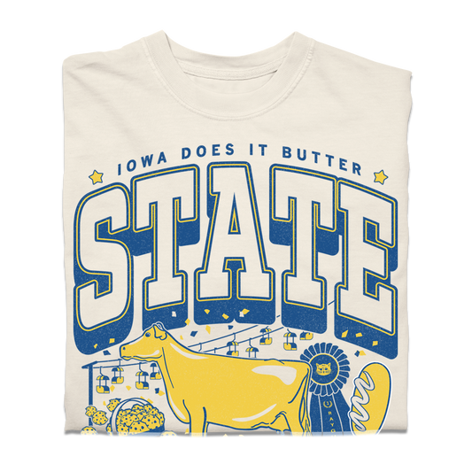 A heavyweight cream-colored t-shirt with a graphic that reads IOWA DOES IT BUTTER and STATE in blue lettering, featuring a yellow cow, a blue ribbon, and a basket of food.