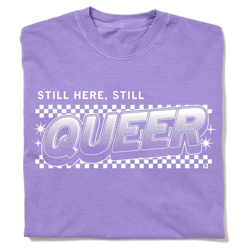 A purple garment-dyed crewneck t-shirt featuring a white STILL HERE, STILL QUEER graphic over a checkered pattern.
