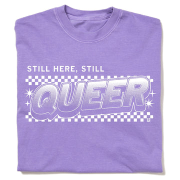 A purple garment-dyed crewneck t-shirt featuring a white STILL HERE, STILL QUEER graphic over a checkered pattern.